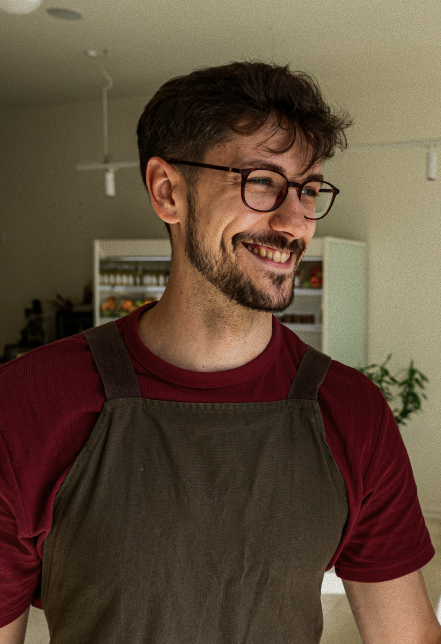 Man with glasses and beard wearing a maroon shirt and brown apron, smiling.